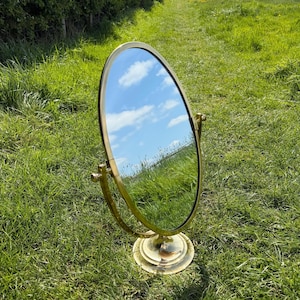 May include: A vintage oval vanity mirror with a brass frame and base. The mirror reflects a blue sky with fluffy white clouds. The mirror is standing on a bed of green grass.