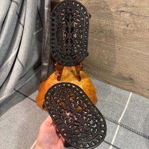 Może przedstawiać: Dwa czarne żeliwne podstawki z misternymi wzorami kwiatowymi. Podstawki mają owalny kształt i podwyższony brzeg. Stoją na drewnianej powierzchni.