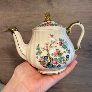 Może przedstawiać: Kremowy ceramiczny imbryk z kwiatowym wzorem w kolorze różowym, niebieskim i zielonym. Imbryk ma złote akcenty na dziobku, uchwycie i pokrywce. Imbryk ma około 15 cm wysokości.