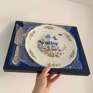 Puede incluir: Juego de plato y servidor de pastel Aynsley en una caja azul marino. El plato blanco tiene un borde festoneado y un diseño floral con el logotipo de Aynsley. El servidor tiene un patrón floral a juego.
