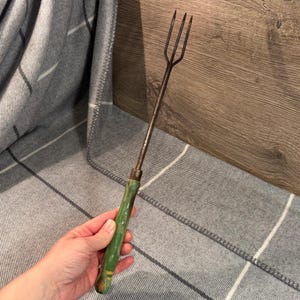 May include: A vintage metal roasting fork with a three-pronged head and a green painted wooden handle. The handle has a gold stripe near the base. The fork is designed for cooking over an open fire or in a fireplace.