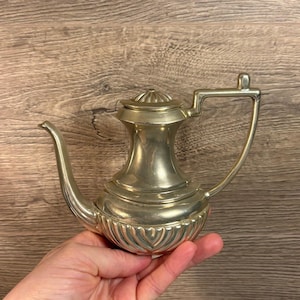 Puede incluir: Una tetera de plata con un asa decorativa y una base acanalada. La tetera es pequeña y tiene un diseño clásico.