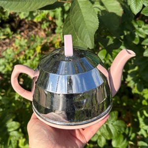 Könnte beinhalten: Dekorative Teekanne mit verspiegeltem, silberfarbenem Körper und rosa Akzenten. Griff, Ausguss und Deckelgriff sind alle rosa. Die Teekanne hat ein einzigartiges, geometrisches Design, ideal zum Servieren von Tee.