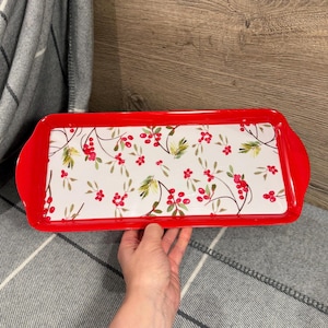 以下が含まれることがあります： 鮮やかな赤い縁とハンドルが付いた長方形のサービングトレイ。トレイの内側は、赤いベリーと緑の葉の繊細な模様で飾られた白い表面です。トレイは手で持たれています。