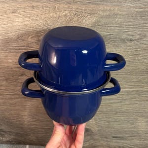 May include: Two cobalt blue enamel pots stacked on top of each other. Each pot has two curved handles and a silver rim. The pots are a deep, glossy blue, and appear to be new.
