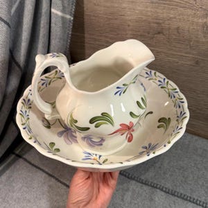 Può includere: Un set di brocca e catino in ceramica bianca. La brocca ha un manico e un beccuccio curvi e si trova all'interno del catino. Entrambi i pezzi sono decorati con disegni floreali nei toni del viola, verde e rosa.