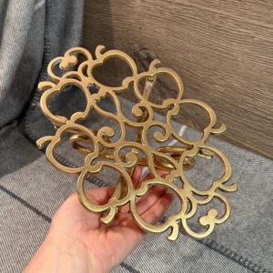 Op de afbeelding: Een goudkleurig metalen decoratief object met een opengewerkt ontwerp met appelvormige elementen. Het object heeft een cirkelvormige basis en wordt in een hand gehouden. De achtergrond is een grijs-witte gestreepte stof.