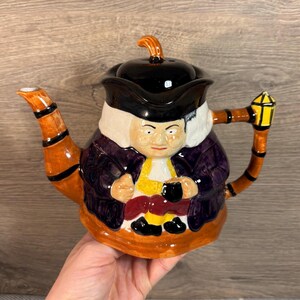 May include: A ceramic teapot shaped like a man wearing a black hat and purple coat. The teapot has a brown handle and spout with black stripes. The man is holding a cup in his hands.