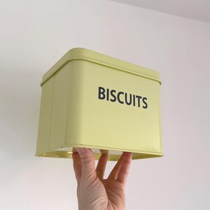 Peut inclure: Une boîte à biscuits rectangulaire jaune pâle avec le mot "BISCUITS" imprimé en noir. La boîte est tenue par une main sur un fond blanc. La boîte a un dessus et un dessous légèrement arrondis.