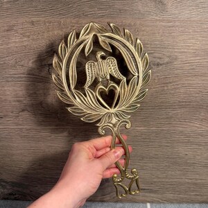May include: A brass trivet with an eagle and heart design. The trivet features a circular wreath with an eagle at the centre, above a heart shape. The handle has a decorative design. The trivet is held by a person.
