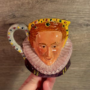 May include: A ceramic pitcher featuring a detailed depiction of a historical figure's face. The pitcher has a yellow crown, a ruffled collar, and a handle adorned with colorful dots. The face is painted with realistic features and warm tones.