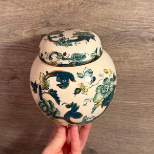 May include: A round, cream-coloured ceramic jar with a matching lid, featuring a decorative pattern of dark teal, green, and yellow floral motifs and swirling leaf designs across the entire surface of the vessel and its rounded lid.