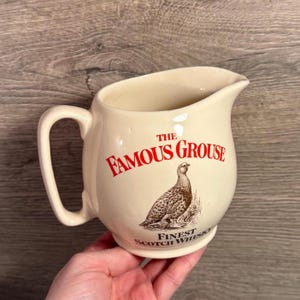 Puede incluir: Jarra de cerámica color crema con asa y pico vertedor. La parte delantera presenta el texto rojo "The Famous Grouse" sobre una ilustración de un pájaro y el texto "Finest Scotch Whisky".