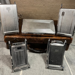 May include: A collection of vintage silver cigarette cases and lighters. The cases and lighters have textured patterns and rectangular shapes. They are arranged on a brown leather case and a gray and white striped fabric.