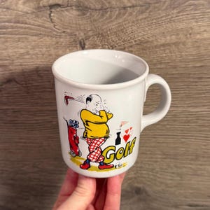 May include: White ceramic mug with a golf-themed cartoon illustration. The design features a golfer in a yellow shirt and red plaid pants, swinging a club. A red golf bag and the word "Golf" are also visible.
