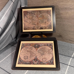 May include: Set of rectangular coasters featuring antique world map designs. Each coaster has a black border and a cream-colored background. The maps are detailed with various illustrations and text, showcasing a vintage aesthetic. The coasters are stacked and displayed on a wooden surface.
