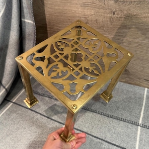 Puede incluir: Un soporte decorativo cuadrado de color latón con un diseño calado en la parte superior. El soporte tiene cuatro patas cuadradas y probablemente se utiliza para exhibir objetos. La parte superior tiene un intrincado patrón floral.