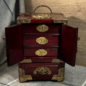 May include: A small, open, wooden jewellery box with a deep red finish. The box has four drawers and two side doors. The corners and edges are trimmed with brass-coloured metal. A brass handle is on top.