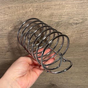 Puede incluir: Un servilletero de metal cromado brillante. El soporte tiene un diseño en espiral con una base redondeada y un asa curva. El soporte está diseñado para guardar servilletas y se muestra sobre un fondo de madera.