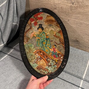 May include: Oval-shaped wall art with a black frame, featuring a bas-relief scene of a woman in a green kimono and a child on a bridge. The artwork includes a red flowering tree and a traditional building.