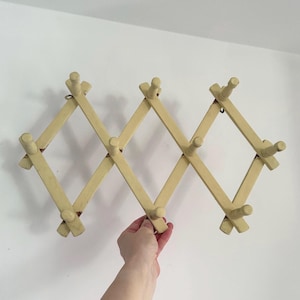 May include: A vintage, expandable wooden coat rack with a light yellow finish. The rack features multiple pegs for hanging items and a diamond-shaped design. The rack is held against a white wall.