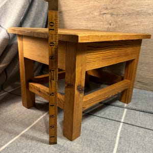 May include: A small, wooden step stool with a rectangular top and four sturdy legs. The stool is made of light-colored wood and measures approximately 10 inches in height. A vintage measuring tape is placed next to the stool.