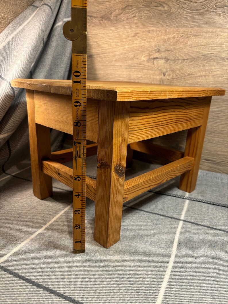 May include: A small, wooden step stool with a rectangular top and four sturdy legs. The stool is made of light-colored wood and measures approximately 10 inches in height. A vintage measuring tape is placed next to the stool.