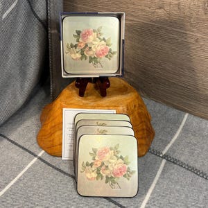 Könnte beinhalten: Ein Set quadratischer Untersetzer mit einem Blumendesign mit rosa und cremefarbenen Rosen. Die Untersetzer befinden sich in einer Schachtel und sind gestapelt, einige werden separat angezeigt. Der Hintergrund ist hellgrün mit einem silbernen Rand.