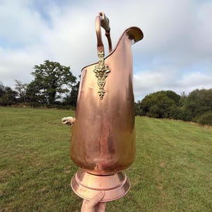 Peut inclure: Une grande houillère en cuivre antique avec un emblème et une poignée en laiton décoratifs. La houillère a une base ronde et un bec verseur. La poignée est en cuivre et en bois. La houillère est tenue devant un champ vert et un ciel nuageux.