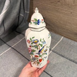 May include: A rustic wooden sign with the text "Welcome" in a handwritten style. The sign is painted white with a distressed finish, and features a decorative floral element in the corner. The background is a blurred image of a wooden surface.