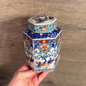 May include: A hexagonal ceramic jar with a removable lid, featuring a white base decorated with blue, orange, and green patterns. The design includes detailed illustrations of figures among trees, floral motifs, and geometric borders on the lid.