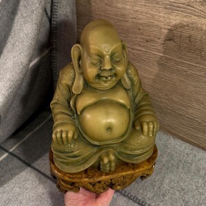 May include: A seated Buddha statue with a serene expression. The statue is a muted green color and sits on a decorative wooden base. The Buddha is depicted with a large belly and detailed robes.