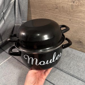 Puede incluir: Dos ollas de esmalte negro apiladas una encima de la otra. La olla inferior tiene la palabra "Moules" en escritura blanca. Ambas ollas tienen asas negras y un borde plateado. Las ollas están sobre una manta gris.
