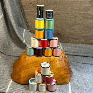 Könnte beinhalten: Eine Sammlung bunter Garnrollen, die auf einem Holzsockel gestapelt sind. Die Spulen sind in verschiedenen Farben gehalten, darunter Gelb, Blau, Grün, Rot und Rosa. Das Garn ist mit dem Markennamen "Drima" und anderem Text beschriftet.