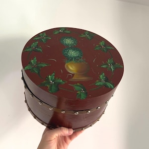 May include: A round, wooden box with a dark red finish. The lid features a painted design of green foliage and a gold-colored vase with green plants. The box is accented with metal studs around the sides.