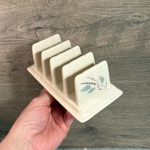 Puede incluir: Tostador de cerámica color crema con cuatro ranuras. El estante presenta un diseño decorativo de hojas azules y doradas. El estante está sostenido por una mano sobre un fondo de madera. Un artículo de cocina vintage.