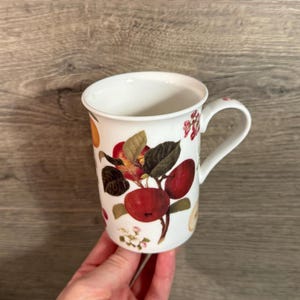 Könnte beinhalten: Weiße Keramiktasse mit einem botanischen Design aus roten Äpfeln, grünen Blättern und kleinen weißen Blüten. Die Tasse hat einen gebogenen Henkel und einen leicht ausgestellten Rand. Das Design ist auf der Außenseite aufgedruckt.