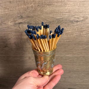 May include: A decorative brass cup filled with wooden matches. The matches have blue tips and are arranged upright. The cup has an ornate design and is held in a hand.