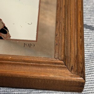 May include: A rectangular wooden frame with a mirrored surface. The frame is a warm brown color with visible wood grain. The text "1919" is visible on the mirrored surface. The frame is set against a textured, light-colored background.