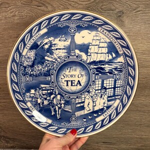 Peut inclure: Une assiette décorative bleue et blanche avec un bord doré, représentant l'histoire du thé. L'assiette présente des illustrations de la cueillette, du transport, de la livraison et de la dégustation du thé, avec le texte "The Story of Tea".