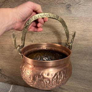 May include: A copper pot with a decorative embossed design and a brass handle. The handle has a unique design with a triangular pattern.