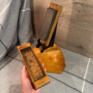 May include: A wooden knife sharpening stone with a dark grey stone and a wooden base. A wooden box with a textured interior is held in the foreground. The wooden base is on a wooden stump.