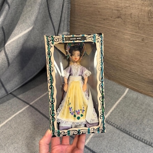 Può includere: Bambola Devon Maid vintage nella sua scatola originale. La bambola indossa un vestito lavanda e giallo con dettagli floreali. La scatola ha un bordo decorativo e il testo "Devon Maid".