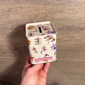 May include: A white metal money box with a circus-themed pattern featuring acrobats, tents, and performers. The box has a slot on top for inserting money and the brand name "Emma Bridgewater" is printed on the front.