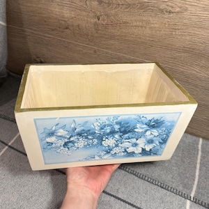 May include: Rectangular wooden planter box with a light cream interior and a blue floral design on the front. The box has a gold trim around the top edge and is held by a hand.