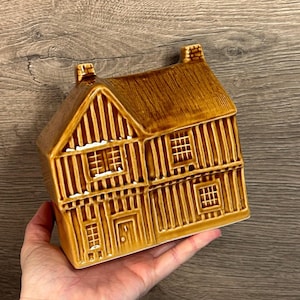 May include: A brown ceramic house ornament with a thatched roof and detailed facade. The house features multiple windows and a door, with vertical and horizontal lines creating a textured look. The ornament is held in a hand.