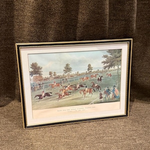 Könnte beinhalten: Ein gerahmter Druck mit dem Titel Race for the Great Ebor St. Leger 1859, der ein Pferderennen mit Jockeys auf einer grünen Bahn zeigt, umgeben von Zuschauern und Bäumen. Das Kunstwerk befindet sich in einem schwarz-goldenen Rahmen mit weißem Passepartout.