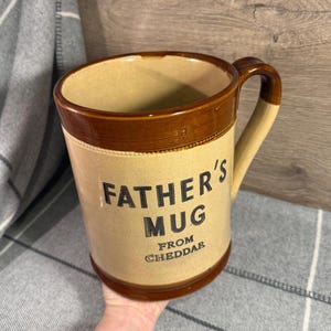 May include: A large, tan ceramic mug with a brown rim, handle, and base. The mug features the text "FATHER'S MUG" in black lettering, with "FROM CHEDDAR" below. The mug is cylindrical with a slightly flared top.