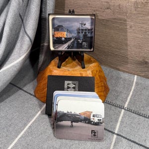 Puede incluir: Un pequeño cuadro con una foto de un tren y un barco sobre un soporte de madera, con un juego de posavasos con imágenes de vehículos de transporte. Los posavasos tienen el texto "Serving the Services".