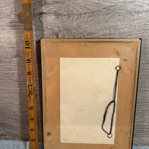 May include: The back of a framed picture with a brown paper backing. A vintage folding ruler is next to the frame. The frame is rectangular and has a black border. The ruler is made of wood and brass and measures up to 14 inches.